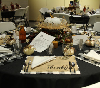 A white ceramic pumpkin with "welcome" written in gold rests on a bed of straw and is surrounded by other pumpkins and candles while the place settings feature black and white checked placemats with some topped with burlap placemats that say "thankful" for Setting the Tables. 
