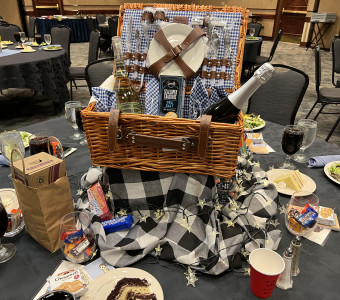 A wicker picnic basket sits on a black and white checked cloth in the middle of the table, with place settings, wine, crackers, lights and more inside and around it for Setting the Tables. 