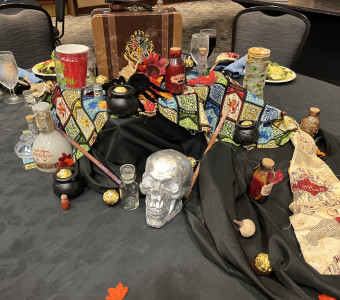 A piece of colorful Harry Potter themed fabric is covered with various potions in small bottles, chocolates, wands, pumpkins and leaves, and a silver skull while a Harry Potter metal lunch box perches on top for Setting the Tables.
