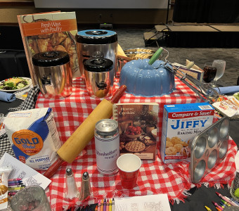 A red and white checked tablecloth sits in the middle of a table, topped with vintage bakeware and cookbooks, including storage canisters, a bundt pan, a manual hand mixer, a rolling pin, a muffin tin and more for Setting the Tables.