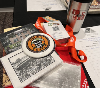 A Lyon County History Center silent auction lot, including a silver LCHS branded travel mug, sticker, red lanyard, button, playing cards, books, postcards and a t shirt.