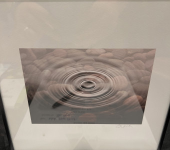 A black framed, white matted black and white photograph of concentric water rings over pebbles in the background.