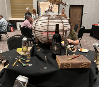 An airship perches atop a sphere filled with cotton clouds and red light, and it surrounded by wine, coins, a leather bound book and more as a table centerpiece. 