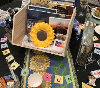 A wooden box filled with Kansas items including a sunflower, books, postcards, and a puzzle sits surrounded by a banner reading "Kansas fun" with bags of chips and candies made in Kansas as a table centerpiece.