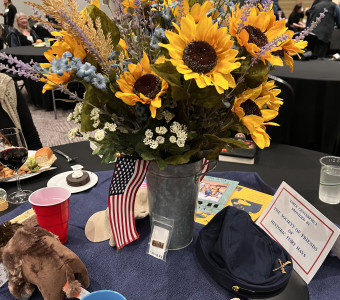A galvanized vase filled with a beautiful arrangement of artificial flowers featuring sunflowers, surrounded by a stuffed bixon, a cavalry hat and pin, mugs and more as a table centerpiece.