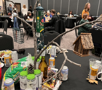 A branch with birdfeeders and birdhouses hanging off of it is surrounded by birdfood, wine, beer, and mugs as a table centerpiece.