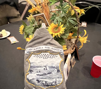 A basket with artificial sunflowers, wheat, and postcards is shown with an "Ad Astra Per Aspera" shirt featuring an image from the seal on the Kansas State flag of a farmer plowing, horses, a house, and a steamboat in the background as a table centerpiece.