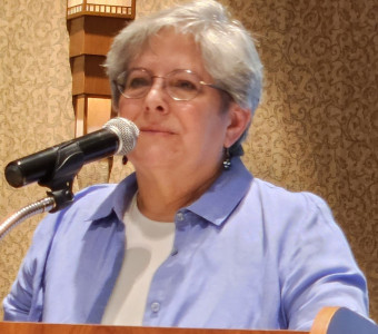 Cori Sherman North speaks into a microphone while wearing glasses, earrings, and a purple collared shirt.