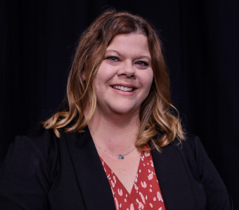 Image of Jamie Buster, wearing a red printed blouse and a black jacket against a black background.