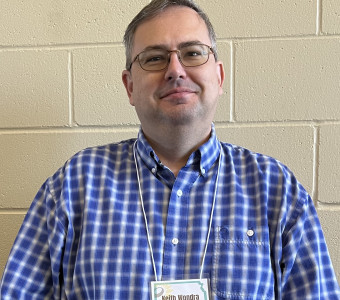 Image of Keith Wondra wearing glasses, a blue and white checked button up shirt, and a nametag on a lanyard.
