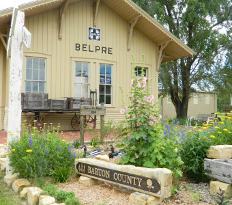 Image of the tan railroad depot with "Belpre" on the side, a wooden wagon and flowers out front around a stone sign that says 'Barton County."