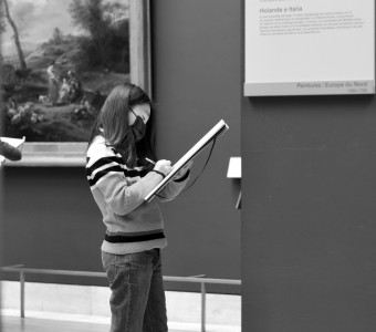 Black and white image of a girl writing on a notepad with a painting behind her and museum label in the foreground by Tommy on Unsplash.      