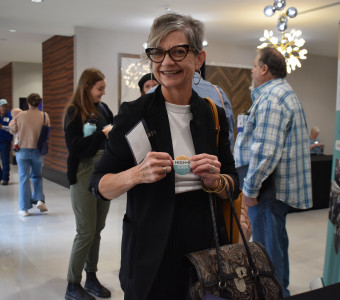A woman dressed in black and white with black glasses, gray hair, and a bag smiles as she holds a High 5 1 Up Club pin.