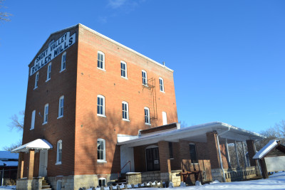 Exterior of the Lindsborg Old Mill & Swedish Heritage Museum.  The 3 story red brick building has "Smoky Vallley Roller Mills" painted in black and white on the side, and the ground is covered in snow. 