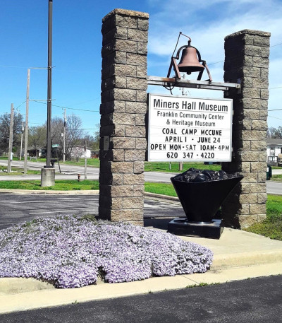 A sign in between two stone pillars is topped with a bell with a coal statue below reads "Miners Hall Museum - Franklin Community Center & Heritage Museum"