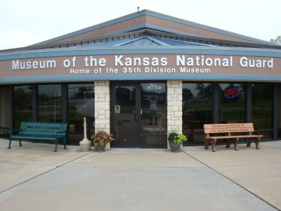 Exterior photo with sign above that reads "Museum of the Kansas National Guard - Home of the 35th Division Museum." Below the sign the glass doors are flanked by two stone pillars and benches.