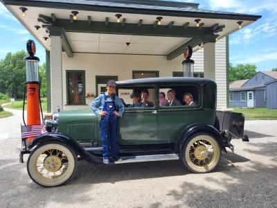 A green and white service station with old-fashioned orange and black Phillips 66 gas pumps sits behind a green vintage car with a young boy in overalls standing on the running board and five other children sitting inside.