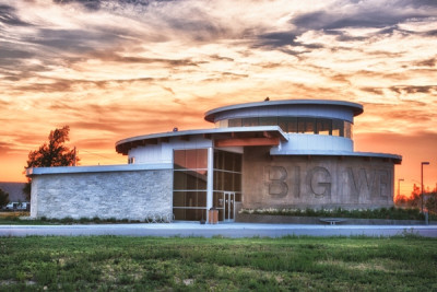 An exterior rendering of the stone and glass Big Well Museum with the name carved in the side in front of a sunset sky