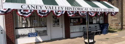 Front of the Caney Valley Historical Society with a black and white striped awning bearing the historical society's name at the front and red, white and blue bunting below shielding large windows and an entry door.