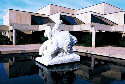 A statue of a horse, a rider, and a bison sits in a pool of reflecting water in front of the concrete and glass Kansas Museum of History