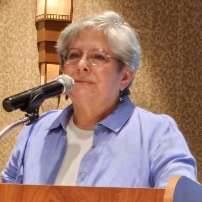 Cori Sherman North speaks into a microphone while wearing glasses, earrings, and a purple collared shirt.