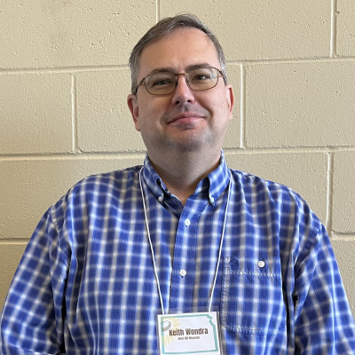Image of Keith Wondra wearing glasses, a blue and white checked button up shirt, and a nametag on a lanyard.