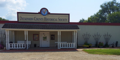 The front of the Dickinson County Historical Society, with a porch roof supported by columns and a sign with "Dickinson County Historical Socity" and a crest of a bison skull and a sunflower above.
