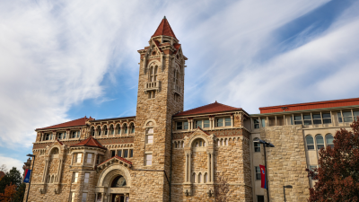 Photo of the stone KU Biodiversity Institute and Natural History Museum with arched windows, a central tower, and red tile roof against a blue sky.