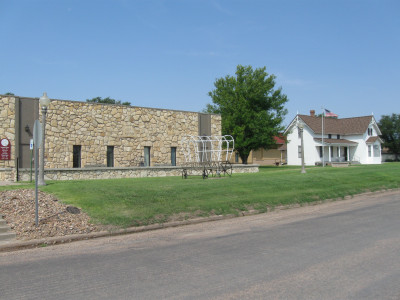 Image of the one story stone Kearny County Museum with windows and a wireframe covered wagon in front next to a 2 story white house with a porch and flagpole with US flag.