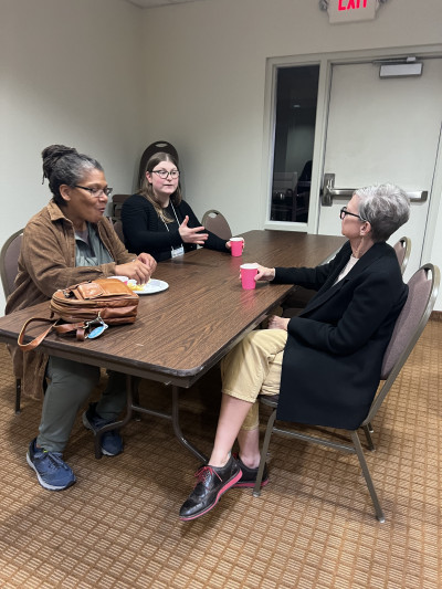 Image of three women sitting at a table having a conversation while two hold red cups and a bag and plate sits in front of the third