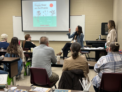 Image of a woman sitting on a desk presenting with a projector screen behind her which reads, "Happy Hour on the Prairie" and people sitting in chairs with their backs to the camera listening.