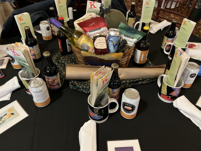 Image of centerpiece with wine, snacks, a hat, and more on a piece of sunflower fabric with a mug, bottle, can, and pamphlets at each table setting from the 2024 Setting the Tables at the KMA Conference in Fort Scott.