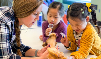 A woman holds a bone as two young Asian girls examine it through magnifying glasses at the Flint Hills Discovery Center