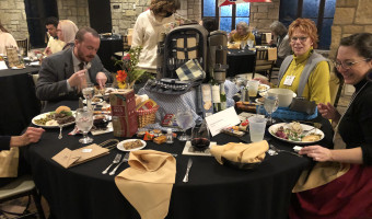 KMA Members sit and stand at a table where a picnic backpack sits open, revealing place settings and napkins and a Wichita city flag while the table features wine, crackers, and cheeses for Setting the Tables.