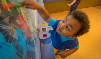 A black child wearing a blue smock fingerpaints on plexiglas at the Kansas Children's Discovery Center.