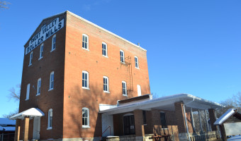 Exterior of the Lindsborg Old Mill & Swedish Heritage Museum.  The 3 story red brick building has "Smoky Vallley Roller Mills" painted in black and white on the side, and the ground is covered in snow. 