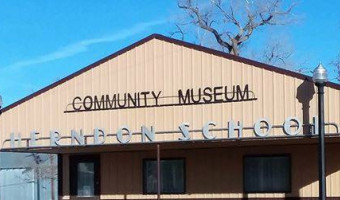 The exterior of a tan metal building with a sign on the front, "Community Museum" and below that "Herndon School"