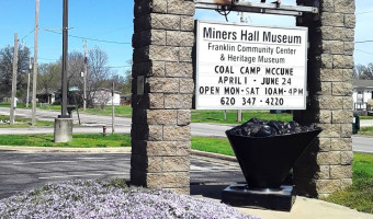 A sign in between two stone pillars is topped with a bell with a coal statue below reads "Miners Hall Museum - Franklin Community Center & Heritage Museum"