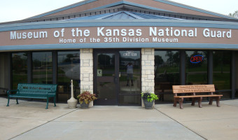 Exterior photo with sign above that reads "Museum of the Kansas National Guard - Home of the 35th Division Museum." Below the sign the glass doors are flanked by two stone pillars and benches.