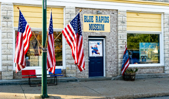 Exterior of of a yellow siding and stone building with a "Blue Rapids Museum" sign in blue above the door and three American Flags on attached to a lightpost near the street.