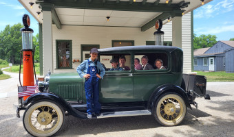 A green and white service station with old-fashioned orange and black Phillips 66 gas pumps sits behind a green vintage car with a young boy in overalls standing on the running board and five other children sitting inside.