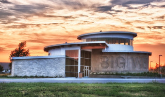 An exterior rendering of the stone and glass Big Well Museum with the name carved in the side in front of a sunset sky