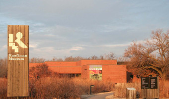 Exterior of the red brick Kauffman Museum with a colorful sign and prairie grass in the foreground