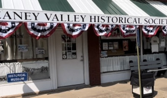 Front of the Caney Valley Historical Society with a black and white striped awning bearing the historical society's name at the front and red, white and blue bunting below shielding large windows and an entry door.