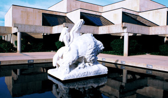 A statue of a horse, a rider, and a bison sits in a pool of reflecting water in front of the concrete and glass Kansas Museum of History