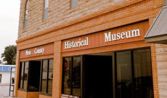 Image of the reddish orange brick and stone facade with "Ness County Historical Museum" in white letters over large glass storefront windows with four arched windows above