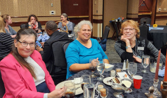 Three women sit at a table with glasses and desserts in front of them.