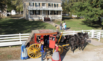 Exterior photo of the two story Mahaffie farmhouse with upper and lower porches and a red stagecoach with yellow wheels pulled by horses and with costumed reenactors is in front.