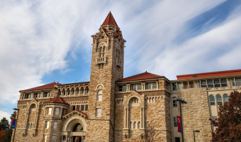 Photo of the stone KU Biodiversity Institute and Natural History Museum with arched windows, a central tower, and red tile roof against a blue sky.