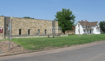Image of the one story stone Kearny County Museum with windows and a wireframe covered wagon in front next to a 2 story white house with a porch and flagpole with US flag.